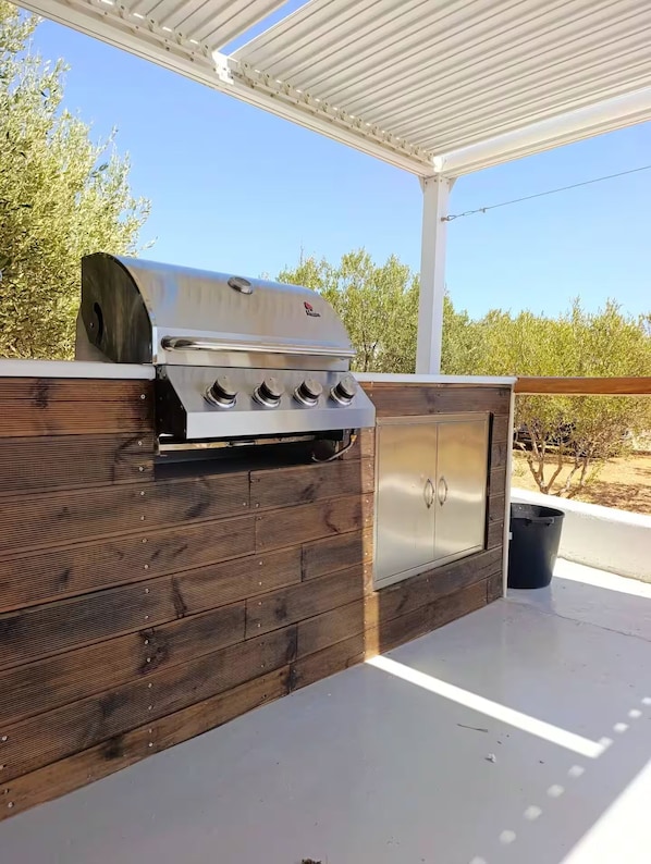 An outdoor grill with a stainless steel finish, a wooden cabinet below, and a covered patio area.