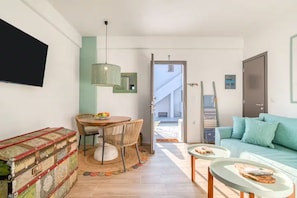 A modern living room with a sofa, a dining table with chairs, a television mounted on the wall, and a view of the outside.