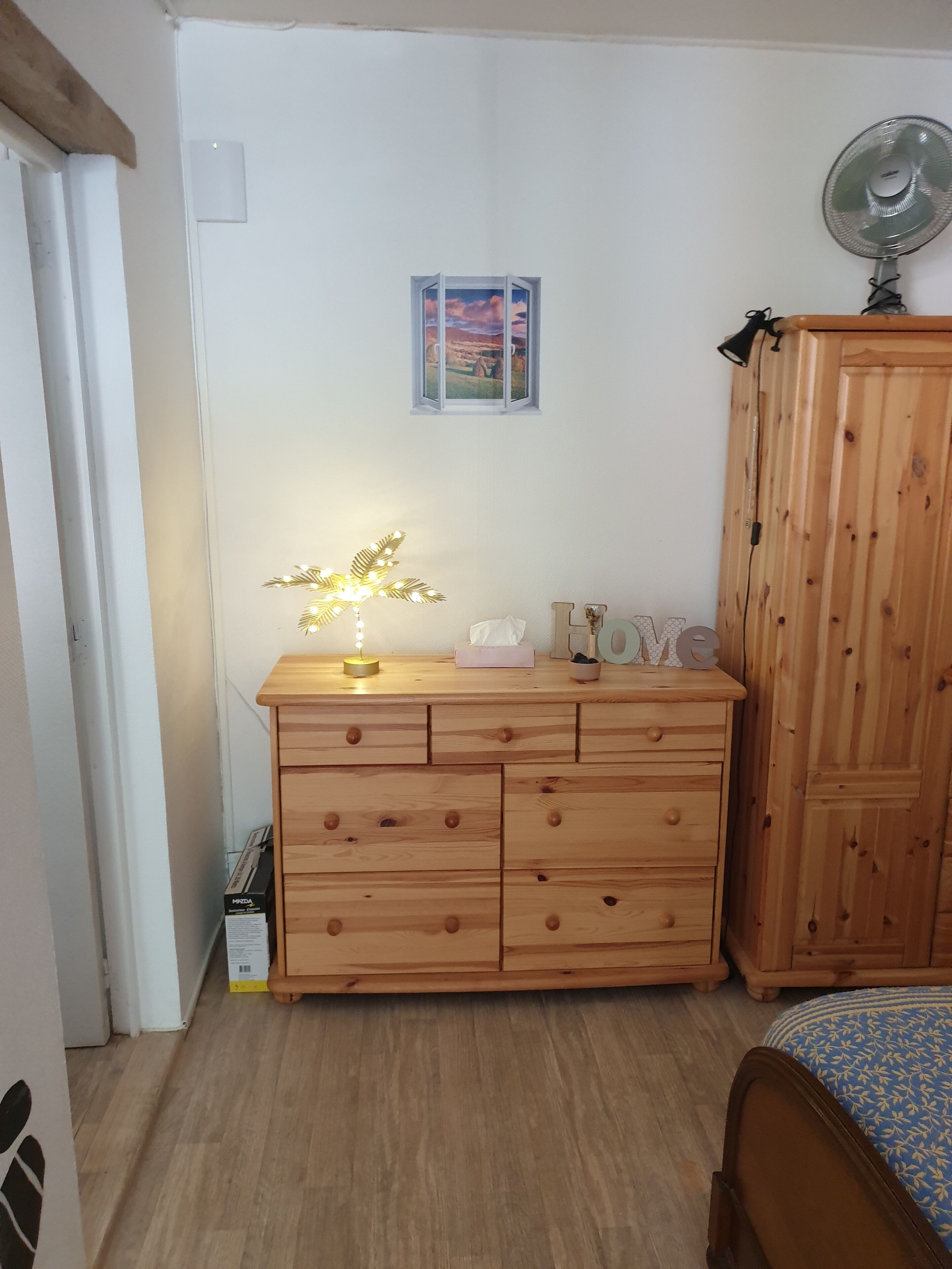 Un dormitorio con cómoda de madera, mesita de noche con lámpara, cama con colcha estampada, armario de madera y una ventana pequeña con vistas al paisaje.