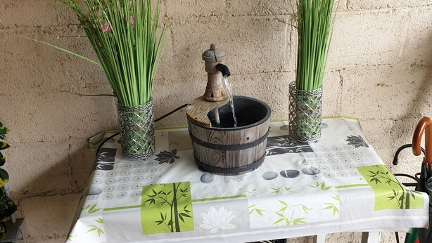 Une table recouverte d’une nappe blanche, deux plantes en pot, une fontaine décorative en bois et un panier placé en dessous.