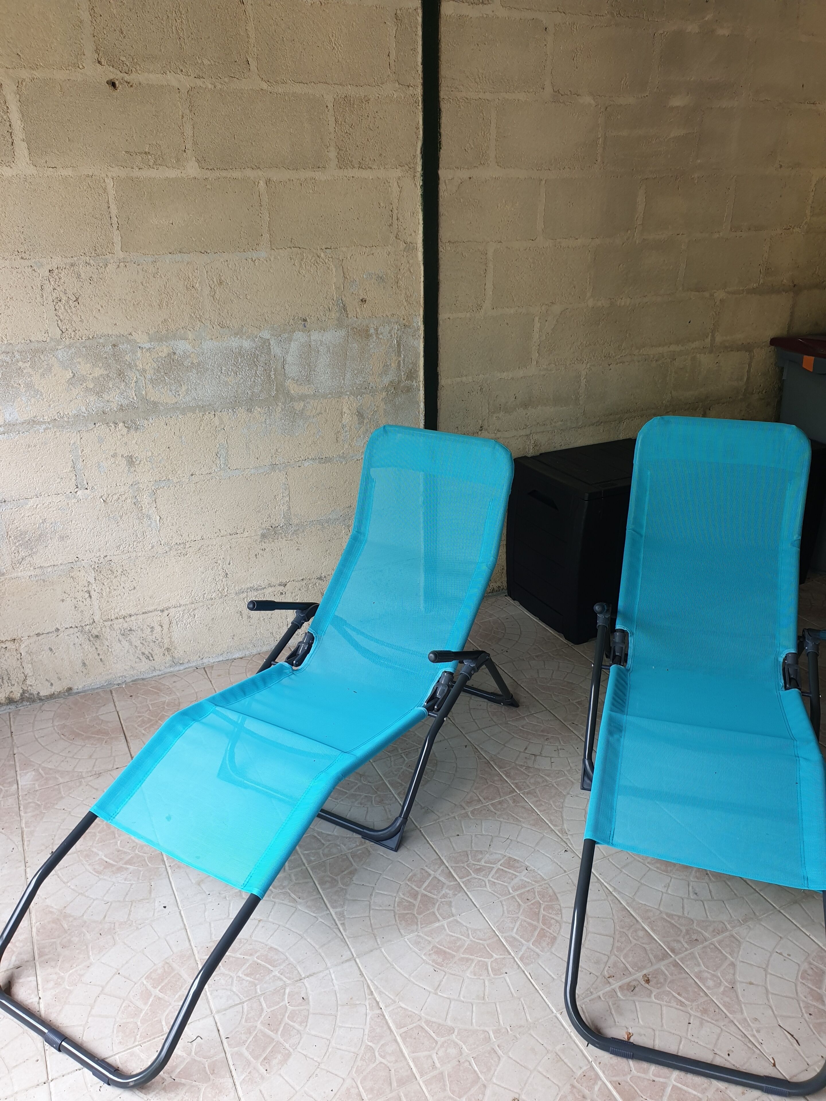 Two turquoise lounge chairs with black metal frames, positioned on a tiled patio against a beige brick wall.
