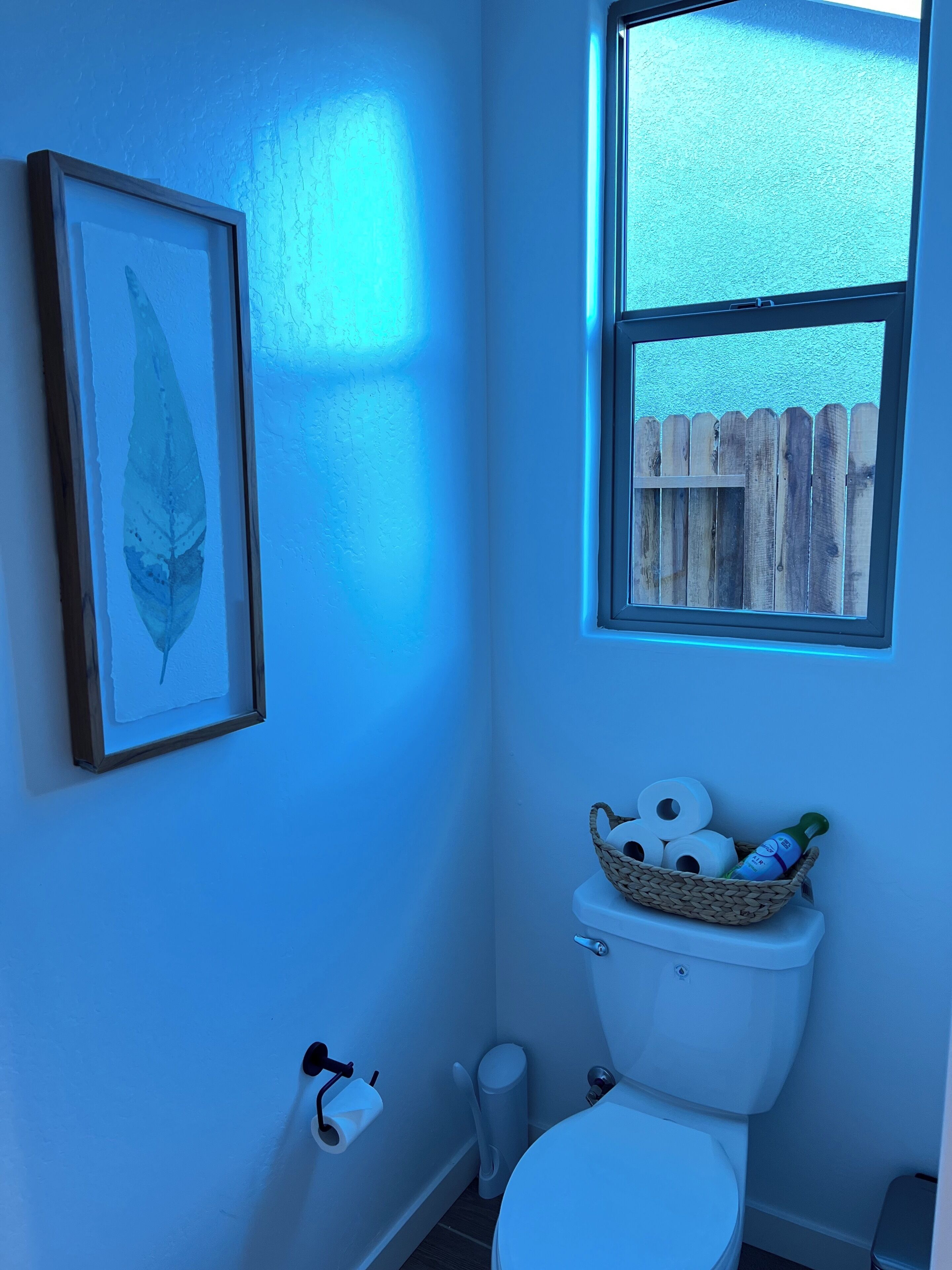 Un baño con inodoro, una obra de arte con una hoja enmarco, una cesta con artículos de aseo y una ventana que da a una valla de madera.