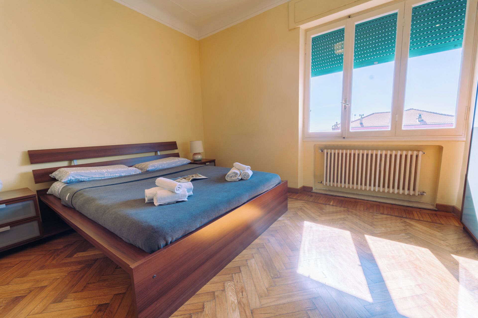 Un lit bien fait, avec des oreillers et des serviettes, une table de chevet avec une lampe, un radiateur et une fenêtre avec des stores.