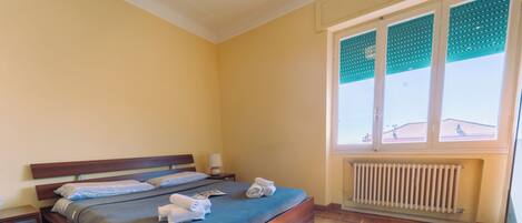 A neatly made bed with pillows and towels, a bedside table with a lamp, a radiator, and a window with blinds.
