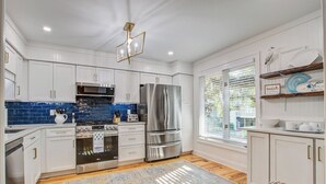 A modern kitchen with white cabinetry, stainless steel appliances, a blue backsplash, and a large window with blinds. - Seabrook Exclusives | 1138 Summerwind (Johns Island)