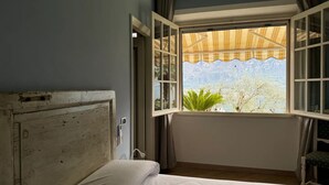 Quarto com cama, janela com cortinas e vista para árvores e montanhas.