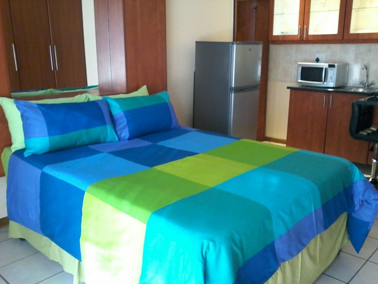 A bedroom with a bed covered in a multicolored bedspread, wooden cabinets, a refrigerator, and a microwave.