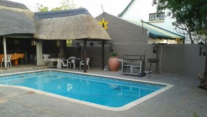 Ein Schwimmbecken mit Ziegelterrasse, Sitzgelegenheiten im Freien und einem strohgedeckten Gebäude.