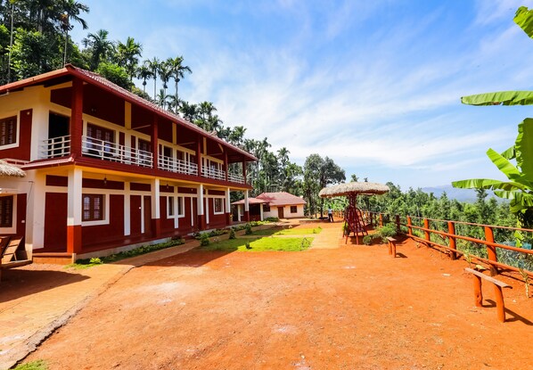 A two-story building with a red and white exterior, a thatched roof, and a paved courtyard with benches and a small garden. - Uttunga Homestay (Mudigere)