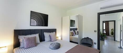 A modern bedroom with a large bed, a headboard, a bedside table, a mirror, and a wall-mounted artwork.
