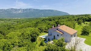 A white house with a red roof surrounded by lush greenery and a mountain in the background.