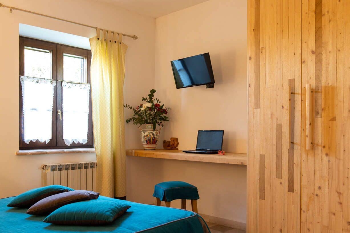 A bedroom with a bed, a desk with a laptop, a TV, and a window with curtains.