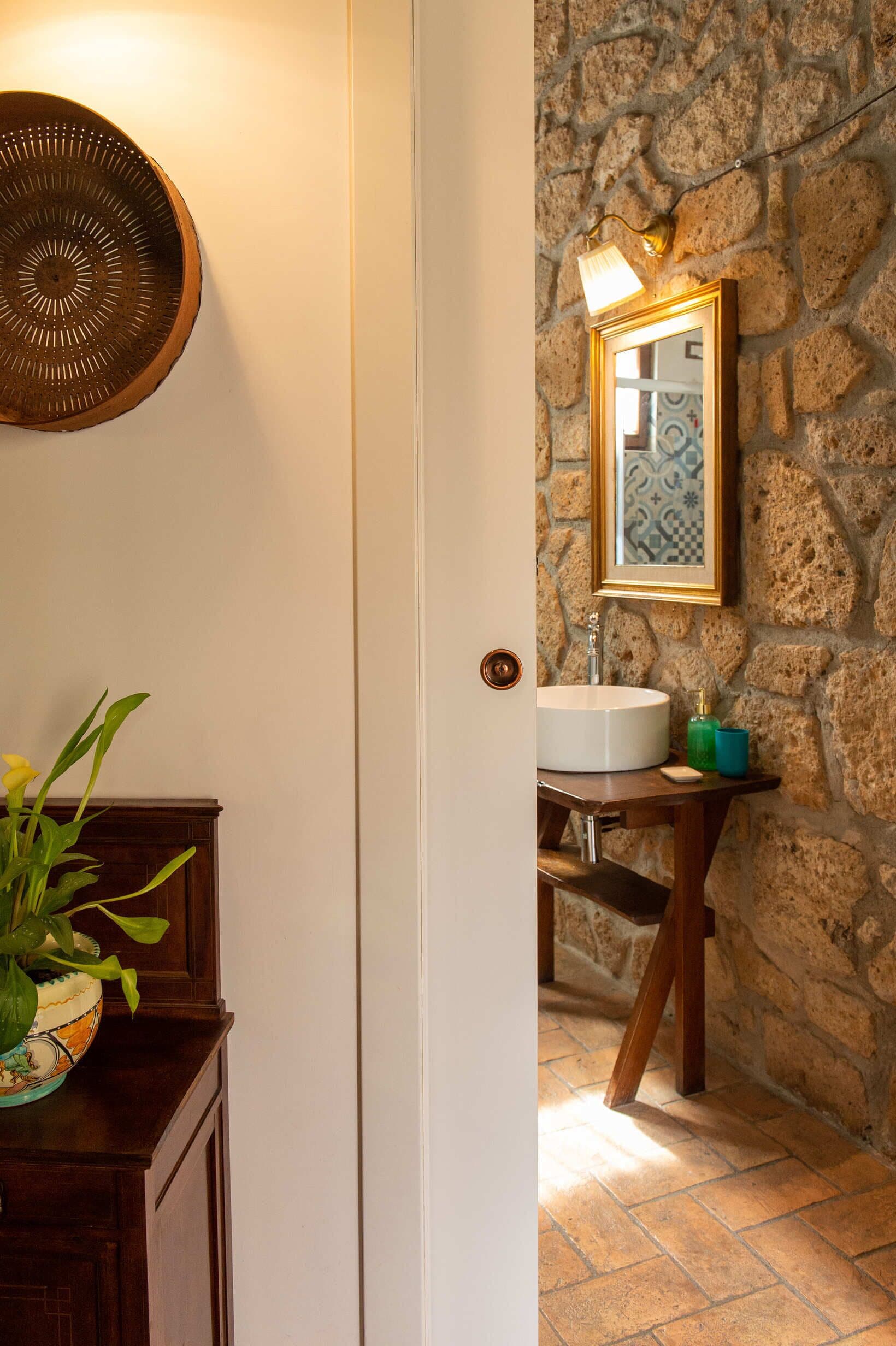 A bathroom with a stone wall, a white sink, a mirror with a gold frame, and a wooden cabinet with a vase of flowers.