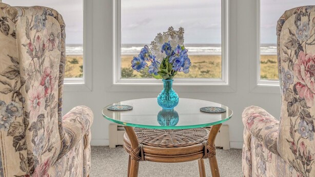 A round glass table with a blue vase of flowers, flanked by two upholstered chairs with floral upholstery, in a room with large windows overlooking a beach. - Beautiful multi-level oceanfront home, perfect retreat for friends or family. (Seaside)