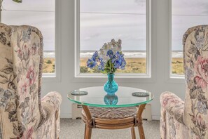 A round glass table with a blue vase of flowers, flanked by two upholstered chairs with floral upholstery, in a room with large windows overlooking a beach. - Beautiful multi-level oceanfront home, perfect retreat for friends or family. (Seaside)