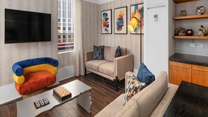 Ein Wohnzimmer mit einer Couch, einem bunten Sessel, einem Couchtisch und einem Fernseher. An der Wand befinden sich gerahmte Kunstwerke und ein Fenster mit Jalousien.