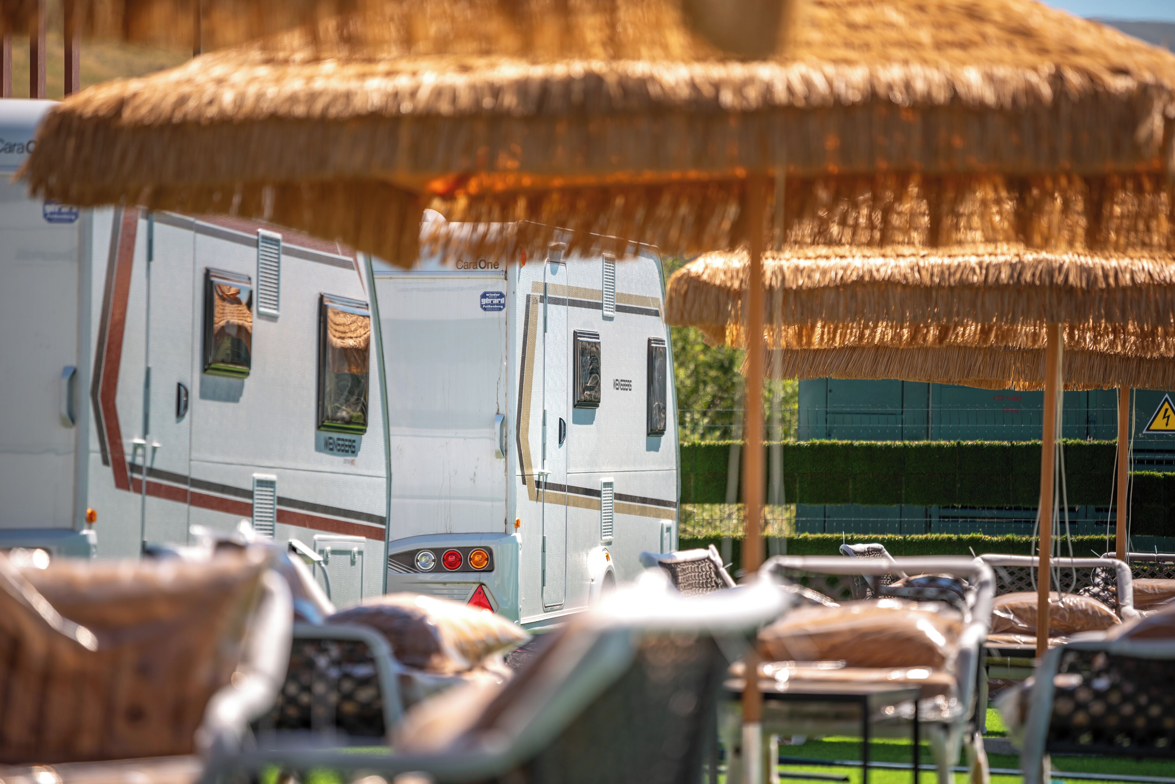 Een rij campers staat onder rieten parasols, met een groene haag en een waarschuwingsbord op de achtergrond.