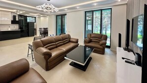 A modern living room with a brown leather sofa, a glass coffee table, and a TV on a white stand. There is a kitchen in the background with a black refrigerator and a dining area. - Comuna Sevan beach Glampings (Artanish)