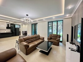 Un salón moderno con un sofá de cuero marrón, una mesa de centro de cristal y un televisor sobre un mueble blanco. Se ve una cocina al fondo con un refrigerador negro y una zona de comedor.