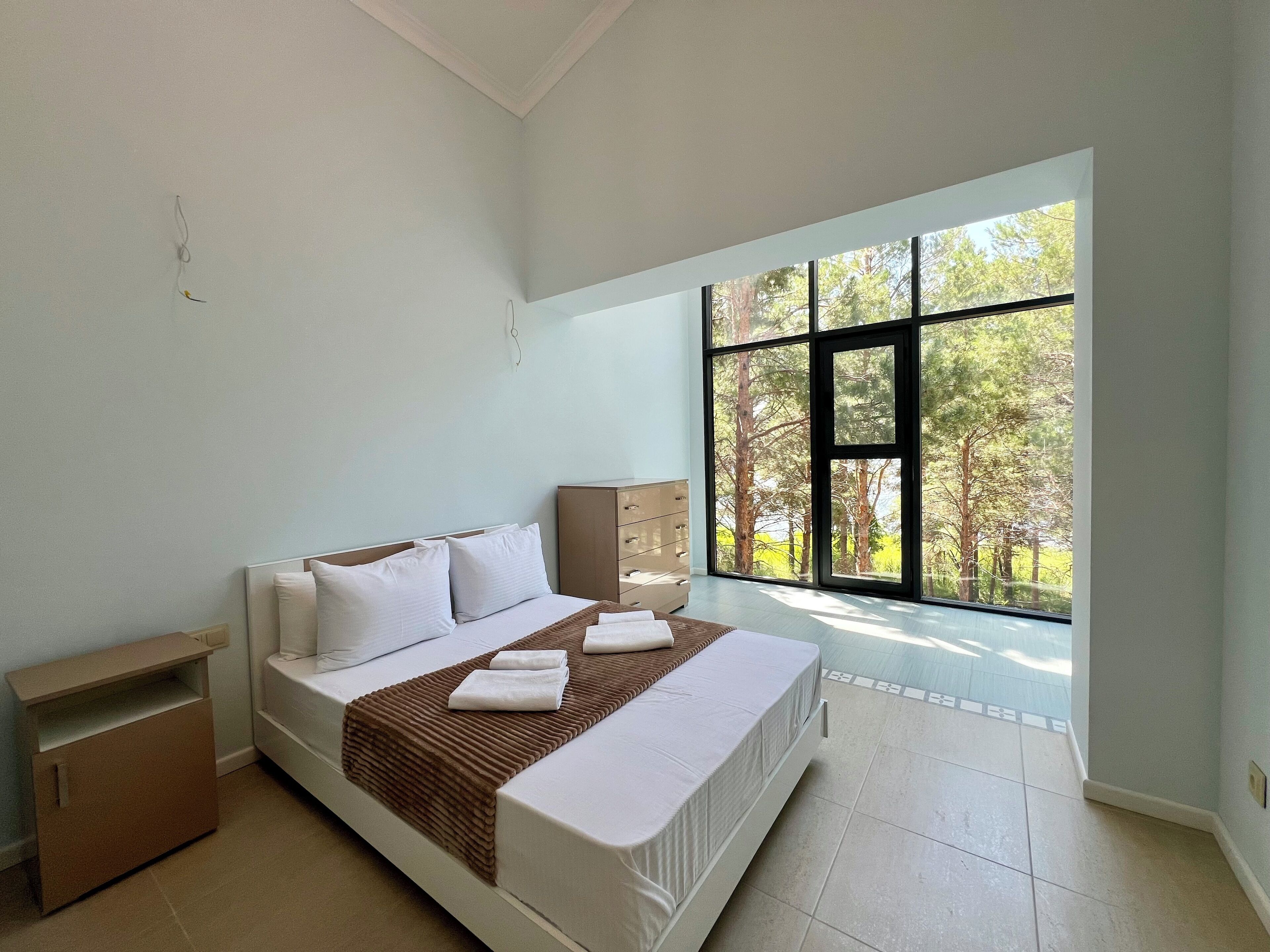 Ein Schlafzimmer mit einem großen Fenster, einem Bett mit weißer Bettwäsche, einem Nachttisch aus Holz und einem kleinen Schrank.