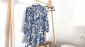Een kamer met een houten kledingrek waarop een blauwe jurk met dessin en een strohoed liggen. Op een zwart plank staat een geweven mand en er is een klein wit tafeltje.