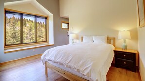 A bedroom with a large window, a bed, a nightstand with a lamp, and a small bedside table.