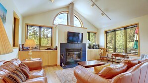 A spacious living room with leather sofas, a fireplace, a television, and large windows offering a view of the outdoors.