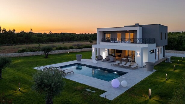 Una casa moderna de dos pisos con piscina, mobiliario exterior y vistas al atardecer.