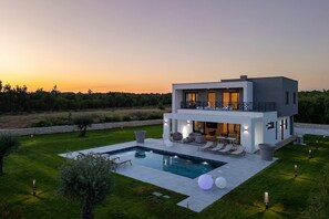 Una casa moderna de dos pisos con piscina, mobiliario exterior y vistas al atardecer.