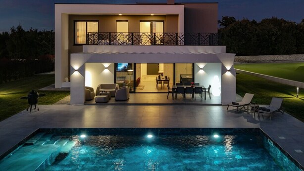 Una casa moderna de dos pisos con piscina, mobiliario exterior e iluminado balcón.