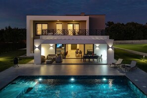 Una casa moderna de dos pisos con piscina, mobiliario exterior e iluminado balcón.