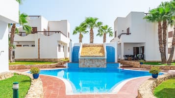 Una piscina con una cascada de piedra, rodeada de edificios blancos y palmeras.