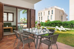 Uma área de refeições ao ar livre com mesa de vidro e cadeiras de vime, porta de correr envidraçada que dá para uma varanda, e vista de um edifício moderno com terraço no telhado.