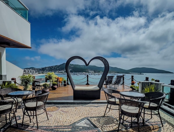 A terrace with outdoor seating, a heart-shaped sculpture, and a view of the sea and hills. - Luxo e Praia Exclusiva para Casal (Arraial do Cabo)