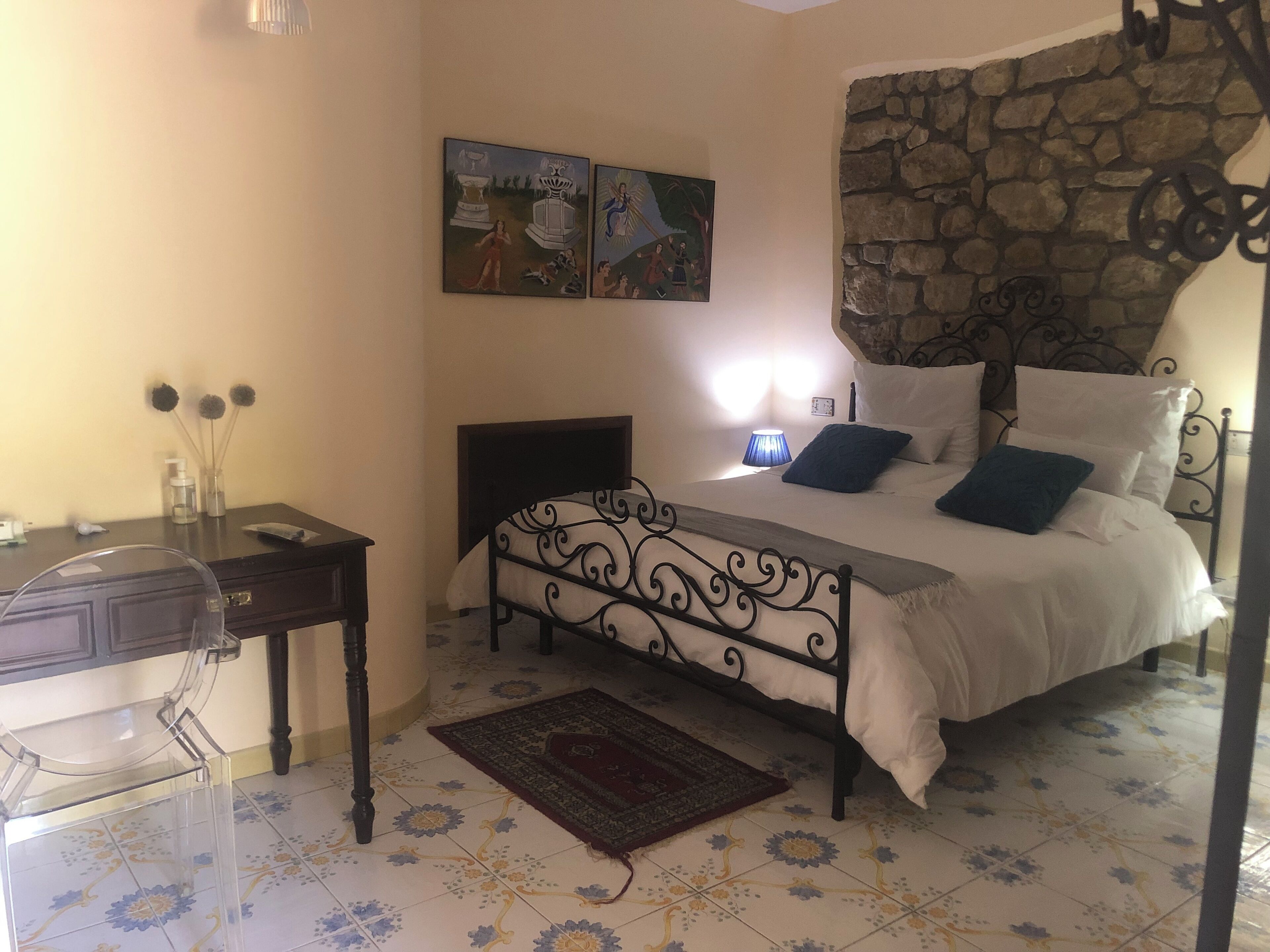 A bedroom with a bed, a desk, and a chair. There are two paintings on the wall, a stone wall, and a patterned floor.