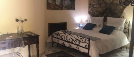 A bedroom with a bed, a desk, and a chair. There are two paintings on the wall, a stone wall, and a patterned floor.