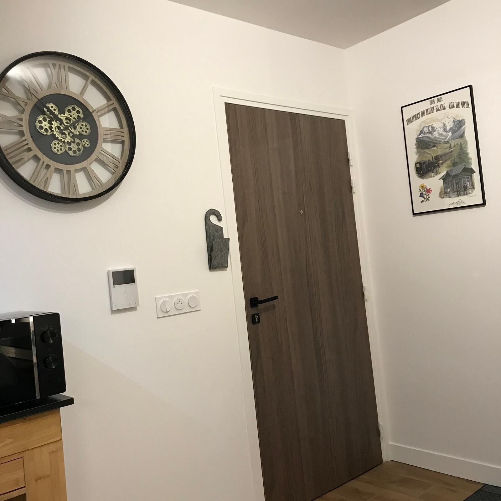 A room with a wooden door, a wall clock, a framed picture, a thermostat, and a small cabinet.