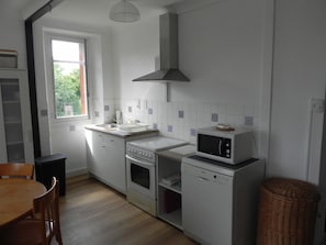 A compact kitchen with white appliances, a window, and a dining area with a wooden table and chairs. - Location Saisonnière Aveyron (Sauveterre-de-Rouergue)