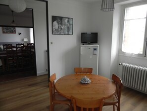 A dining area with a round wooden table and chairs, a white refrigerator with a TV, and a window with blinds. - Location Saisonnière Aveyron (Sauveterre-de-Rouergue)