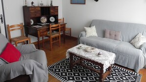 A living room with a grey sofa, two wooden chairs, a small table with a white lace tablecloth, and a patterned rug. - Location Saisonnière Aveyron (Sauveterre-de-Rouergue)