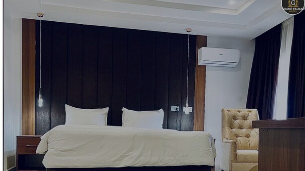 A modern hotel room with a large bed, a wooden nightstand, a chair, and an air conditioning unit on the wall. - Grand Palmac Hotel (Abuja)