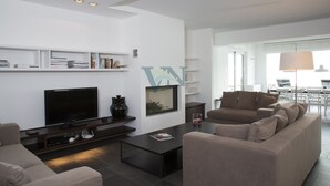 A modern living room with a flat-screen TV, a coffee table, and a sofa. There is a bookshelf with books and decorative items.