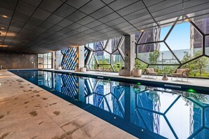 A modern indoor swimming pool with geometric glass walls, lounge chairs, and a view of the cityscape. - Opulent Suite at Westside Place (Melbourne)
