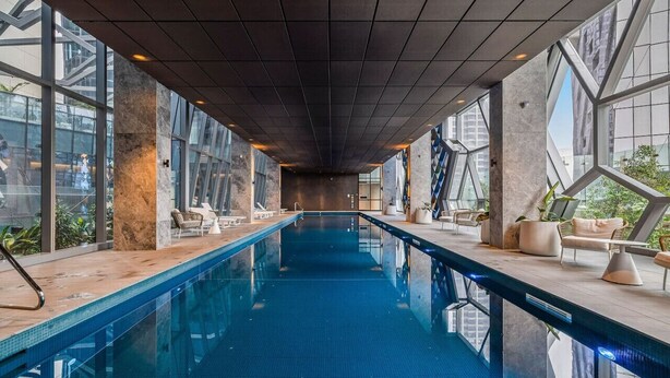 A long indoor swimming pool with clear glass walls, modern seating, and a view of the city.