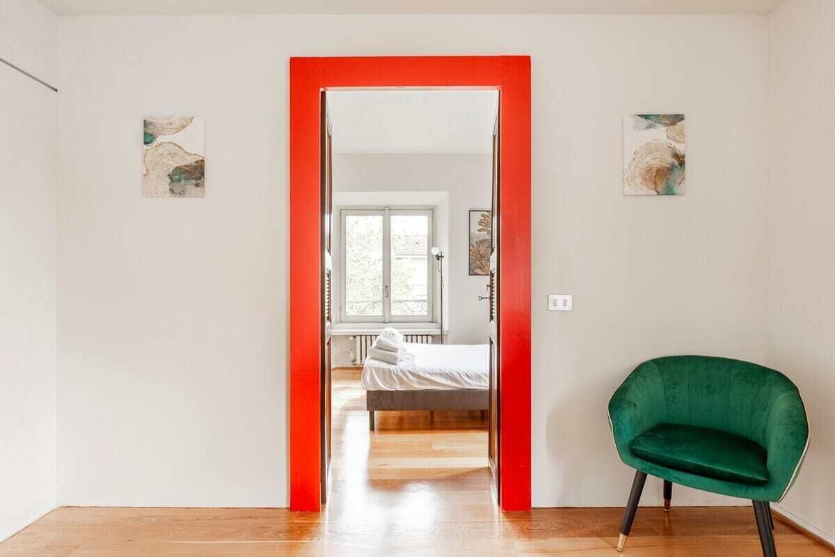 A room with a wooden floor, a green chair, a bed with white bedding, and a bright red door.