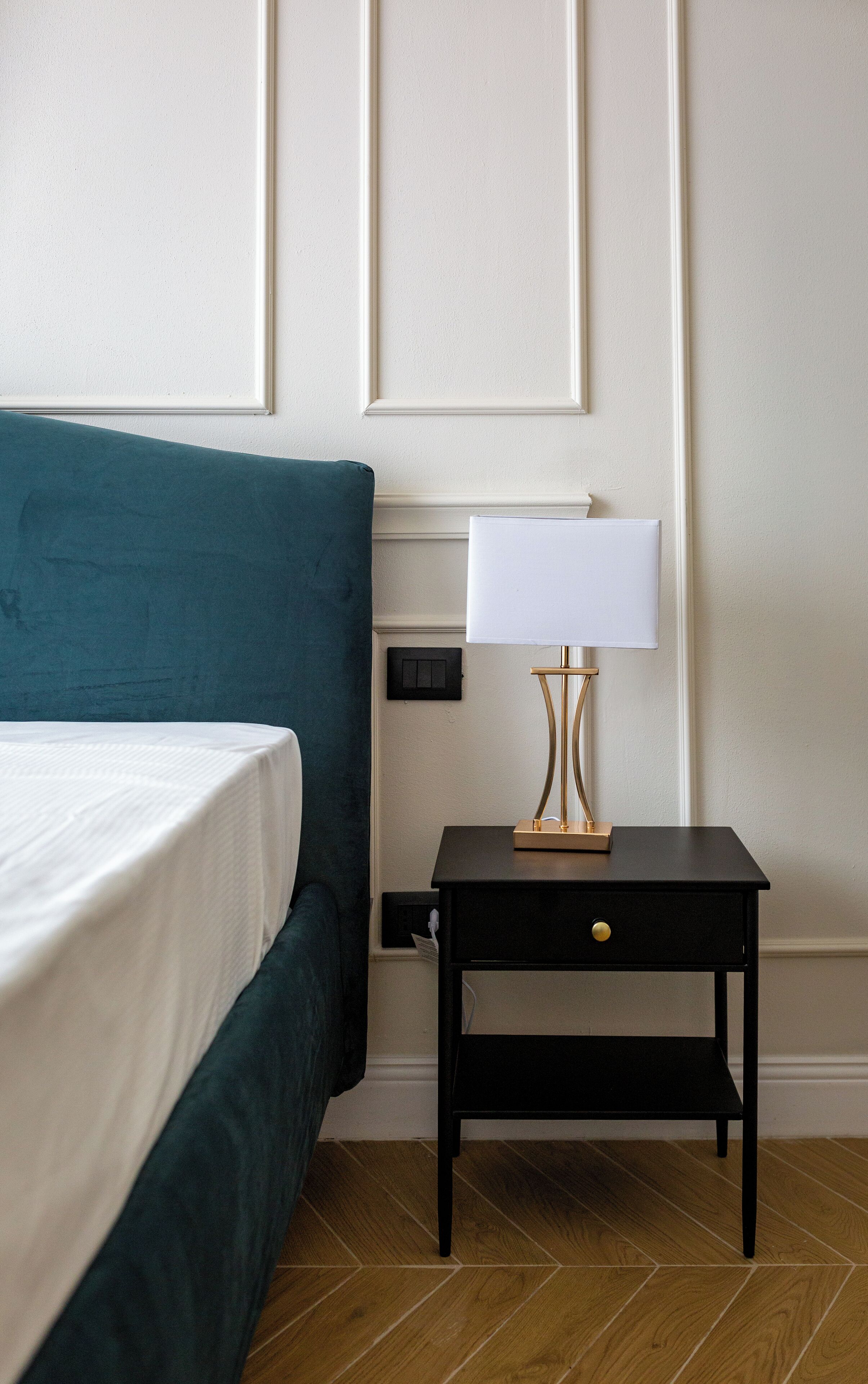 A neatly made bed with a teal headboard, a bedside table with a lamp, and a small drawer.