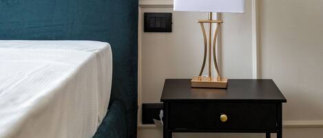 A neatly made bed with a teal headboard, a bedside table with a lamp, and a small drawer.