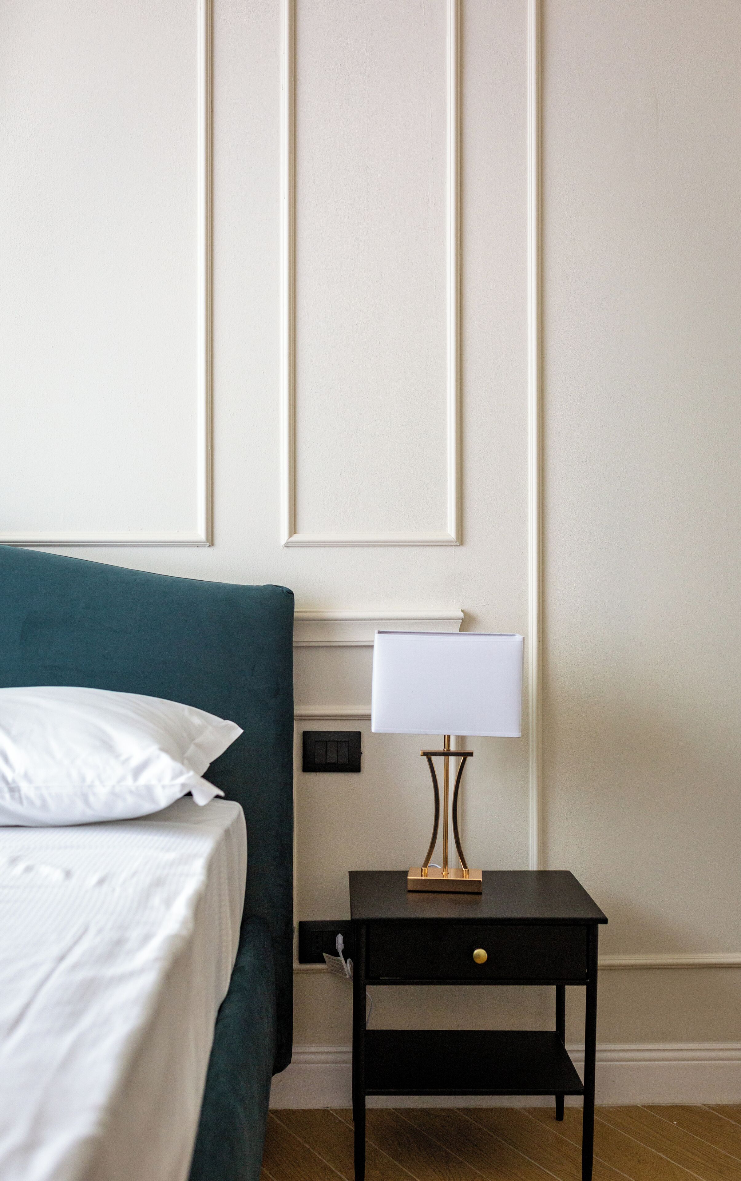 A neatly made bed with white linens and a teal headboard, a bedside table with a lamp, and a wall with paneling.