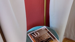 Una revista sobre un pouf redondo color turquesa en una esquina, con una pared roja y una cortina blanca.