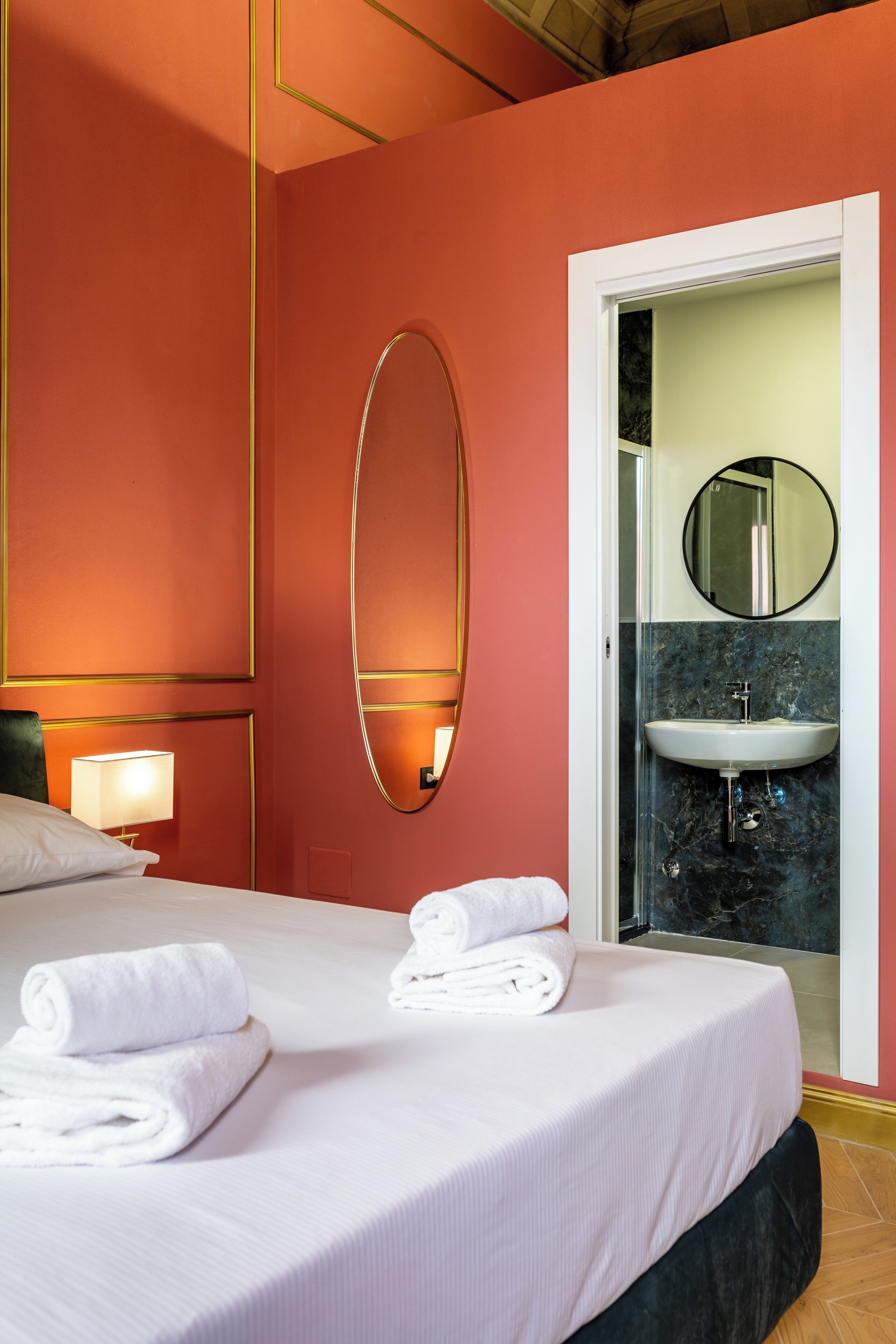 A hotel room with a bed, a large mirror, a bedside lamp, and a bathroom with a sink and mirror.